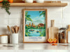 a framed poster of a tropical landscape with boats on a lake, surrounded by palm trees and other natural elements, placed on a kitchen counter.