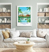 a living room with a large framed poster of a tropical landscape hanging on the wall, depicting a village with palm trees and a river. The poster is surrounded by a white sofa, a coffee table, and a bookshelf filled with books and decorative items.