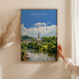 A framed painting of a picturesque town with a river and a church spire in the background, hanging on a wall.