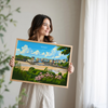 A woman is holding a framed picture of a coastal town, with a beach, buildings, and flowers visible in the image.