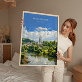A woman holds up a framed painting of a scenic landscape with a river, trees, and a church spire.