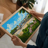 A person is holding a framed print of a colorful illustration depicting a cityscape with buildings, trees, and a body of water.