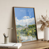 A framed poster of a scenic landscape with a river and buildings, hanging on a wall next to a white vase with dried flowers.