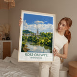 A woman holds up a framed poster of Ross-on-Wye, a town in Herefordshire, England, with a scenic view of the town and its surroundings.